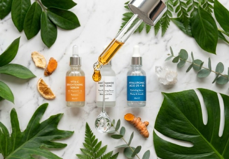 gota de sérum de vitamina C caindo de um conta-gotas com texturas de ácido hialurônico e elementos naturais ao fundo representando ativos cosméticos para a pele