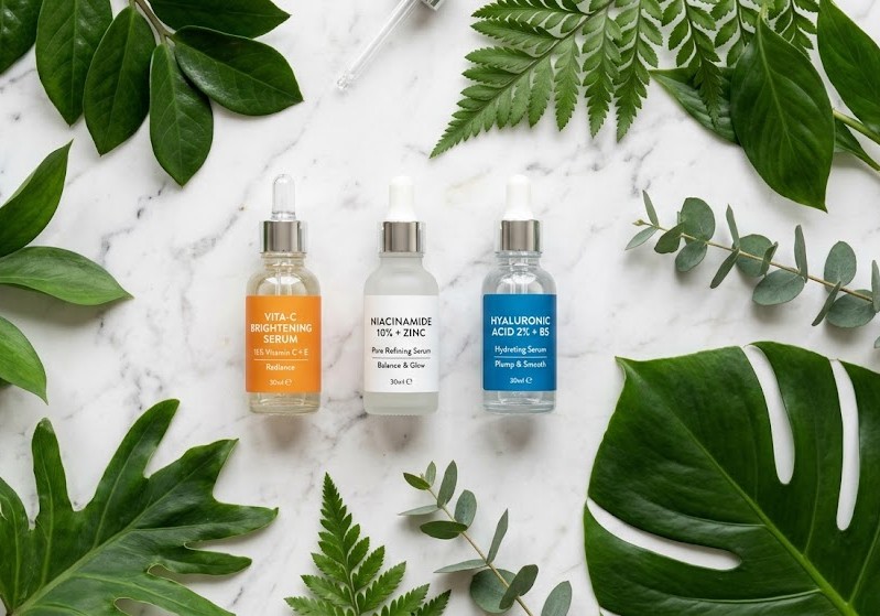 frascos de sérum com vitamina C, niacinamida e ácido hialurônico organizados em flat lay sobre mármore branco com folhas verdes ao redor